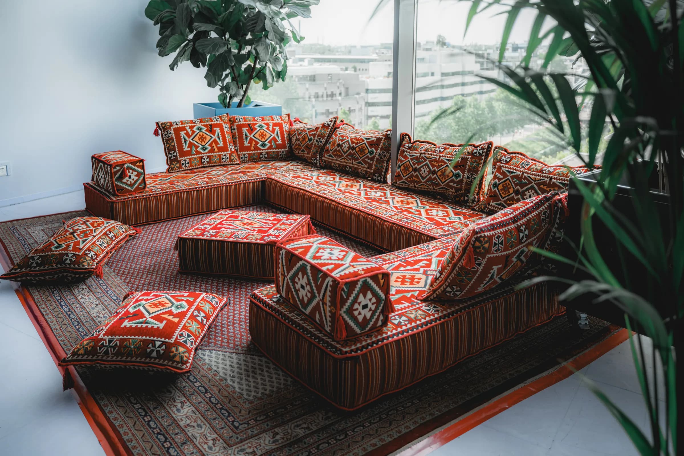 Hoekopstelling van rode kilim-majlisbanken met kussens tussen planten voor een raam